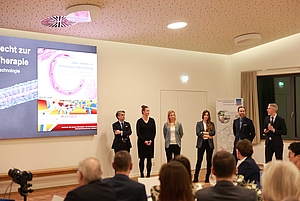 6 Personen stehen vorne und halten einen Vortrag, neben ihnen wird eine Präsentation an die Wand gebeamt.