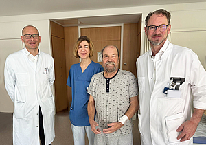 Drei Ärzte (Eine Frau, zwei Männer) in weißem Kittel nebeneinander. Als zweiter von rechts steht ein Patient in grauer kurzer Kleidung in einem Patientenzimmer. 