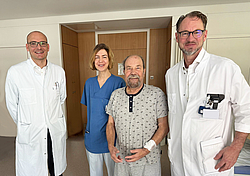 Drei Ärzte (Eine Frau, zwei Männer) in weißem Kittel nebeneinander. Als zweiter von rechts steht ein Patient in grauer kurzer Kleidung in einem Patientenzimmer. 