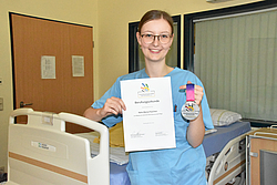 Junge Frau im Kasack vor einem Patientenbett mit Urkunde in der Hand