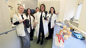 Fünf Frauen im weißen Kittel blicken in die Kamera, in der Hand halten zwei schwarze Figuren, rechts ein Tisch mit Laborutensilien