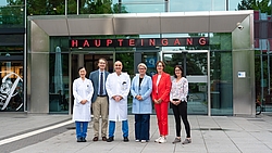Sechs Personen stehen vor einem Gebäude mit der Aufschrift „HAUPTEINGANG“, darunter zwei Männer in weißen Kitteln, ein Mann im Anzug, und drei Frauen in unterschiedlicher Kleidung
