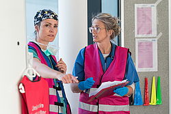 Zwei medizinische Fachkräfte mit Schutzkleidung und farbigen Warnwesten stehen im Gespräch vor einer Wand mit farbigen Kegeln und einem Aushang