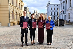 Vier Personen in formeller Kleidung stehen auf einem gepflasterten Platz und halten jeweils eine rote Urkunde vor sich