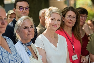 Gruppe von Menschen in einem Raum, im Vordergrund eine lächelnde Frau mit kurzen blonden Haaren und weißem Oberteil