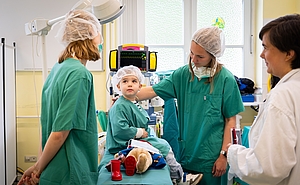Kind sitzt auf OP-Tisch und zwei Ärztinnen in Grün reden mit ihm