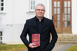 Ein dunkel gekleideter Mann steht mit eine Buch in der Hand vor einem hellen Gebäude.