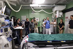Ein Arzt in blauer Arbeitskleidung erklärt einer Gruppe von Personen in einem modernen Krankenhausraum mit medizinischen Geräten und einer Liege.