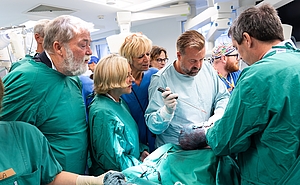 Viele Personen stehen nah beieinander um einen OP-Tisch und schauen gespannt zu.