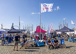 Mehrere Pavillons, Liegestühle und bunte Fahnen sind zu sehen. Menschen sitzen oder stehen in Grüppchen zusammen.