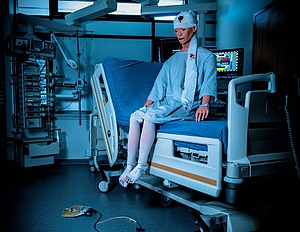 Eine lebensgroße Puppe sitzt auf einem Krankenhausbett. Sie hat eine Verletzung am Kopf und blutet, sie schaut erschrocken und trägt einen Verband der nicht richtig befestigt wurde. Der Raum ist sehr dunkel, im Hintergrund leuchtet das Testbild auf einem Bildschirm hell auf.