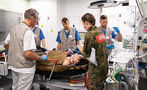 5 Personen in Dienstkleidung, eine davon in Bundeswehruniform stehen an einer Trage um einen Verletzten. Es ist viel Medizintechnik zu sehen.
