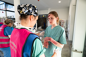 Zwei Personen in medizinischer Schutzkleidung im Gespräch, eine trägt eine bunte Kopfbedeckung und eine pinkfarbene Weste