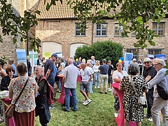 Menschen stehen und unterhalten sich bei einer Veranstaltung im Freien vor einem Backsteingebäude mit Fenstern und begrünten Büschen