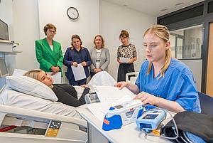Krankenpflegerin in blauer Arbeitskleidung liest Patientendaten neben einer liegenden Patientin im Krankenhausbett, im Hintergrund stehen vier Frauen und beobachten