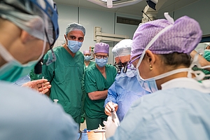 Mehrere Herzchirurgen in Operationskleidung führen eine Operation durch, im Hintergrund beobachten weitere Ärzte die Szene