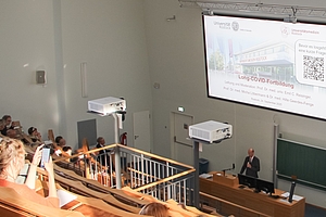 Einige Personen sitzen in einem Hörsaal, man sieht nur etwas von ihren Köpfen. Sie schauen nach vorne zu einer Präsentaion.