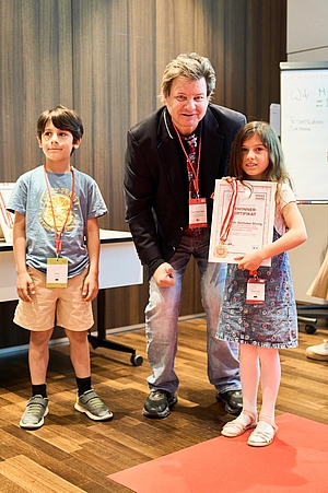 2 Kinder und 1 Erwachsener schauen in die Kamera. Sie tragen alle einen Ausweis. Ein Kind hält eine Auszeichnung in der Hand.
