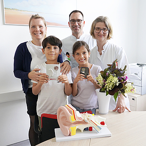 3 Erwachsene und 2 Kinder stehen in einem hellen Arztzimmer und schauen fröhlich in die Kamera.