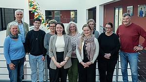 Gruppe von neun Personen steht in einem Innenraum vor einem Geländer und blickt in die Kamera.