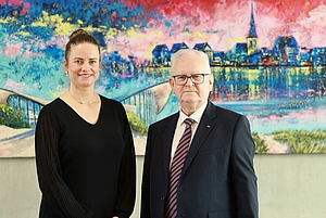 2 Personen stehe lächelnd nebeneinander, hinter ihnen ist eine bunte Wand.