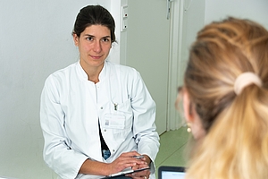 Ärztin im weißen Kittel befragt eine Patientin, die nicht erkennbar im Vordergrund sitzt