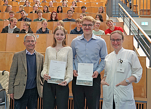 4 Personen stehen lächelnd nebeneinander in einem Hörsaal und 2 von ihnen halten eine Auszeichnung in der Hand.