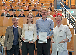 4 Personen stehen lächelnd nebeneinander in einem Hörsaal und 2 von ihnen halten eine Auszeichnung in der Hand.