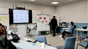 Person steht vor einem Whiteboard mit bunten Haftnotizen und präsentiert vor mehreren sitzenden Personen in einem Besprechungsraum