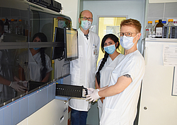 Drei Mitarbeiter in Arbeitskleidung im Labor an einem großen Testgerät 