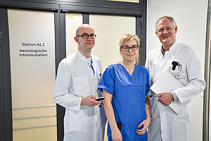 Drei medizinische Fachkräfte in weißen Kitteln und blauer Arbeitskleidung stehen vor einer Tür mit der Aufschrift 'Station A2.2 Neurologische Intensivstation'