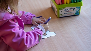 Kind in rosa Jacke malt mit blauem Stift auf ausgeschnittenes Papier auf einem Holztisch