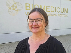 Frau mit Brille und schwarzem Oberteil lächelnd vor einer Wand mit der Aufschrift: BIOMEDICUM UNIVERSITÄTSMEDIZIN ROSTOCK