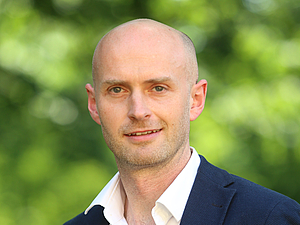 Eine Person mit weißem Hemd und dunklem Sakko vor unscharf grünem Hintergrund