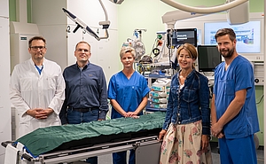 Fünf Personen in unterschiedlicher Krankenhauskleidung und in zivil stehen um eine Trage herum in einem Untersuchungsraum