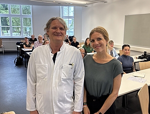 Ein älterer Mann und jüngere Frau in einem Raum, hinter ihnen Sitzen Menschen in Reihen hintereinander