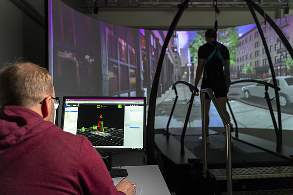 Person sitzt vor einem Bildschirm mit 3D-Ganganalyse-Daten, im Hintergrund steht eine weitere Person auf einem Laufband vor einer großen Projektion einer Straßenszene