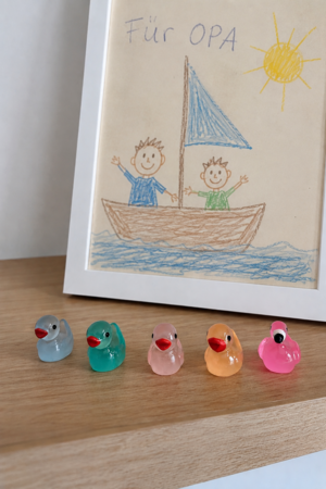 Fünf bunte Gummienten in einer Reihe auf einem Holzregal vor einem Kinderbild mit zwei lachenden Figuren in einem Segelboot und einer Sonne.