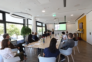 Eine Person hält einen Vortrag vor einer Gruppe von Teilnehmern in einem hellen, modernen Konferenzraum mit großen Fenstern und Holztisch.