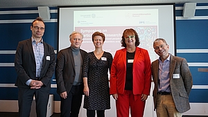 5 Personen stehen nebeneinander vor einer Leinwand und schauen freundlich.