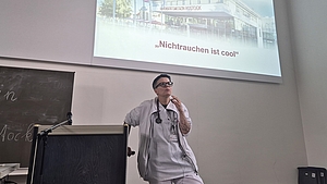 Person in Arztkittel mit Stethoskop steht vor Projektionsleinwand mit Text 'Nichtrauchen ist cool'