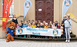 Kinder stehen mit einem Plakat und einem Teddy auf den Stufen vor einem älteren Gebäude