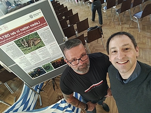 Zwei Männer stehen vor einem Plakat mit einem Bild eines Rehs in einem Raum mit Stuhlreihen