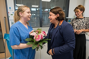 Zwei Frauen lächelnd im Gespräch, eine trägt blaue Arbeitskleidung und hält einen Blumenstrauß, im Hintergrund steht eine dritte Frau mit gemustertem Oberteil.