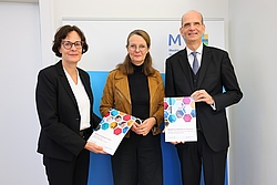 Zwei Frauen und ein Mann stehen vor einer Wand und halten zwei bund Bände zur Medizinstrategie in den Händen.