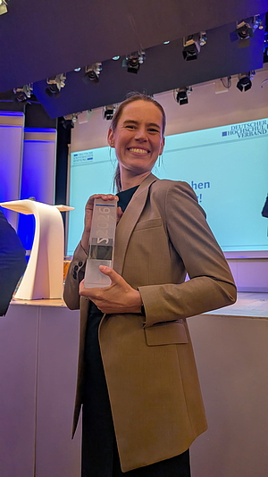 Person in braunem Blazer hält eine gläserne Trophäe vor einer Bühne mit Podium und Bildschirm im Hintergrund