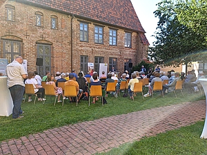 Etwa 20 Menschen sitzen auf Stühlen in einem Garten vor einem historischen Ziegelgebäude und hören einem Redner zu.