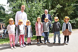 Kinder mit Sonnenhut stehen in Reihe im Grünen, dahinter zwei Ärzte 