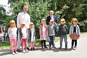 Kinder mit Sonnenhut stehen in Reihe im Grünen, dahinter zwei Ärzte 