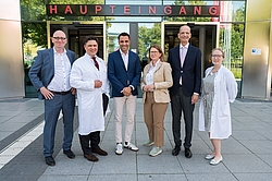 Sechs Personen in formeller und medizinischer Kleidung stehen vor einem Gebäude mit der Aufschrift 'HAUPTEINGANG'